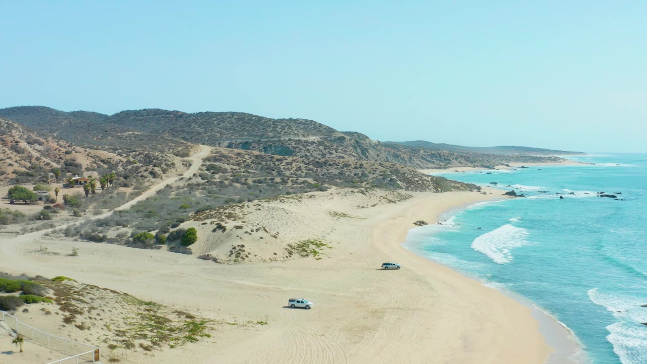 San Luis Beach East Cape Los Cabos is shown with dune scrub in the foreground, two vehicles on the sand, surf to the right, hillside roads behind, and no visible people.