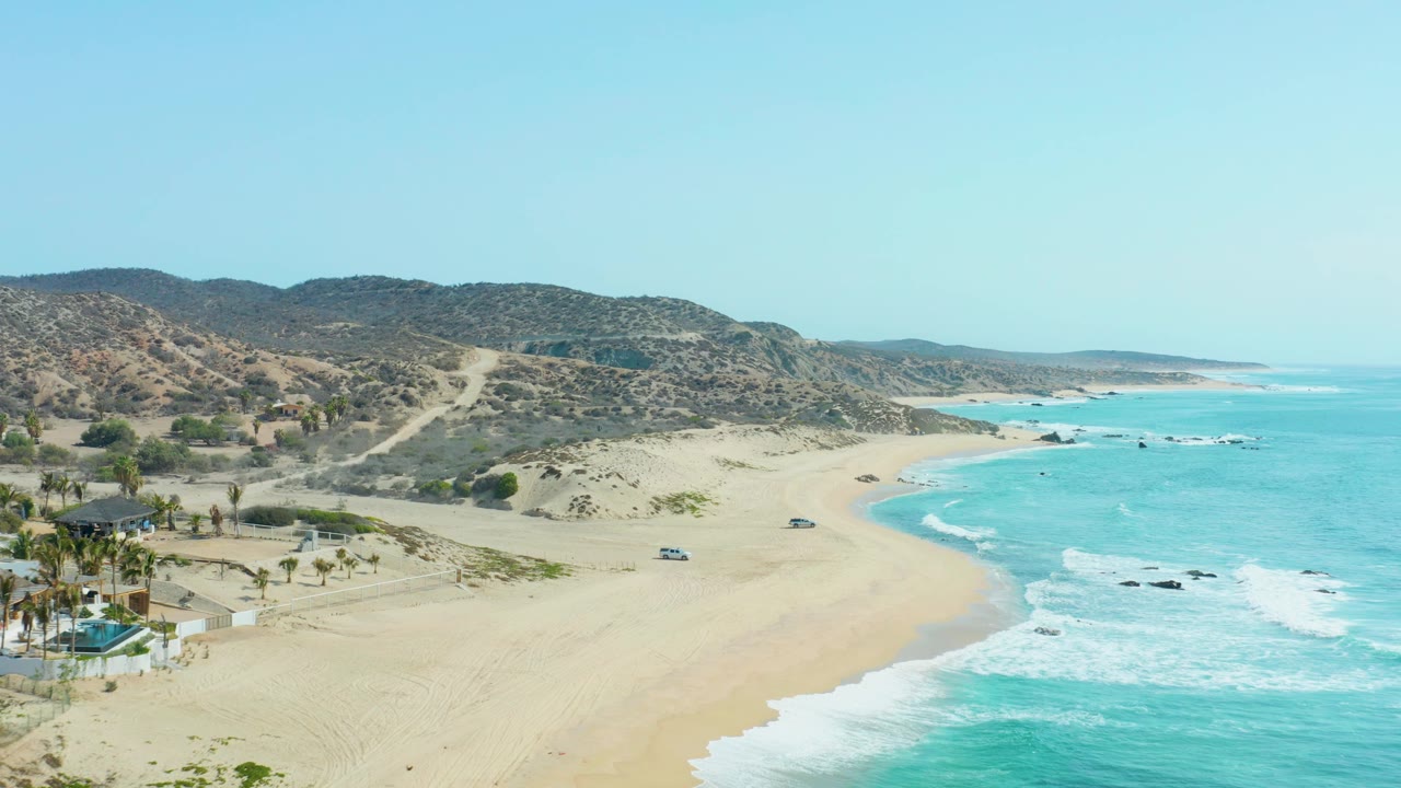 San Luis Beach East Cape Los Cabos seen from above, with pale sand, blue surf, scrub hills, a beach road, two vehicles, and no visible people.