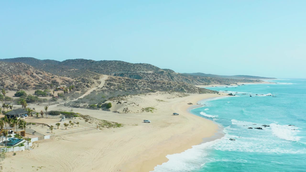 A high aerial frame over San Luis Beach East Cape Los Cabos shows the beach road cutting across open sand, vehicles near the shore, turquoise water, and no visible people.