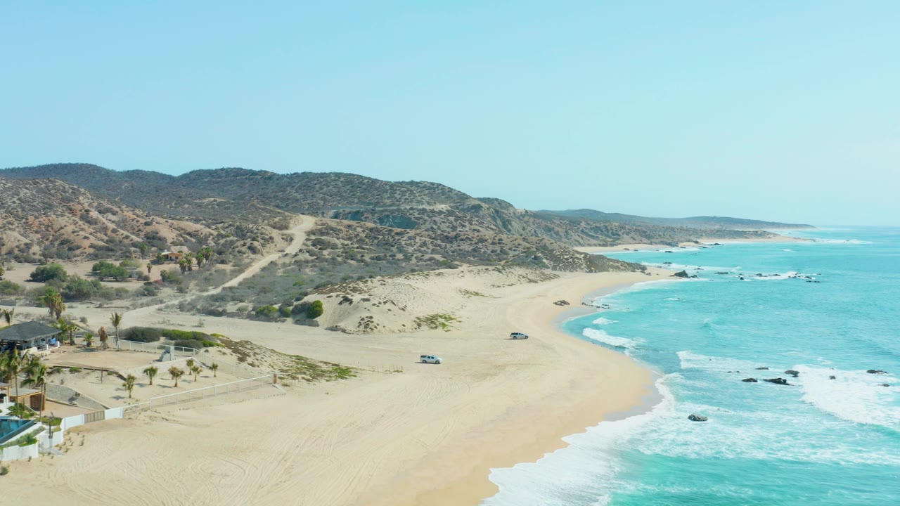 Aerial beach scene with palms, small buildings, road, waves, and hills.