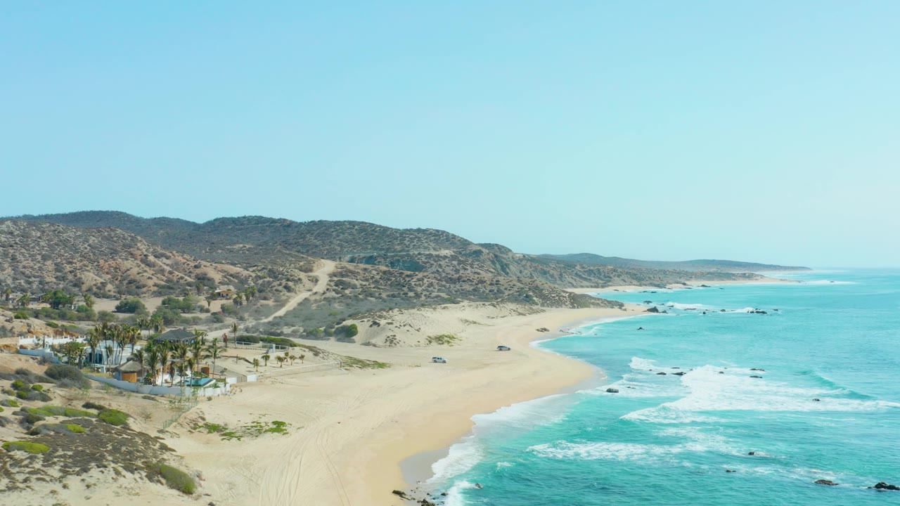 Aerial frame at San Luis Beach East Cape Los Cabos with palm-lined beach structures at lower left, open sand across the center, white surf bands to the right, and no visible people.