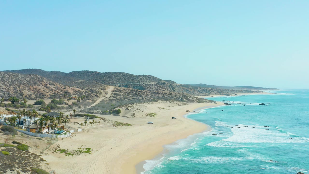 Aerial coastline with beach compound, surf, sand, and hillside road.