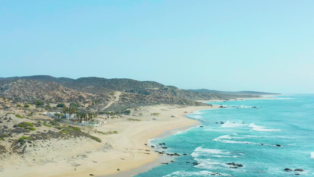 Aerial view of San Luis Beach East Cape with a sandy road at left, turquoise surf at right, low desert hills behind the beach, palm trees near buildings, and no visible people.