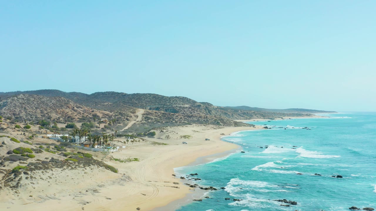Drone perspective of San Luis Beach East Cape with turquoise water filling the right side, tan sand and beach road in the center, scrub-covered hills in back, and no visible people.