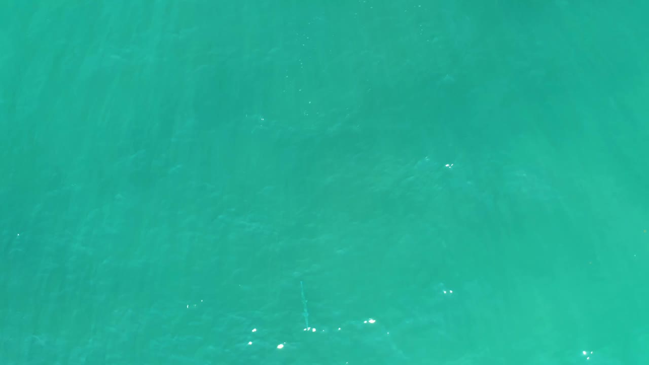 At 0:02, clear blue-green water at San Luis Beach shows small white sun reflections near the surface and diffuse pale shapes below, with no visible people.