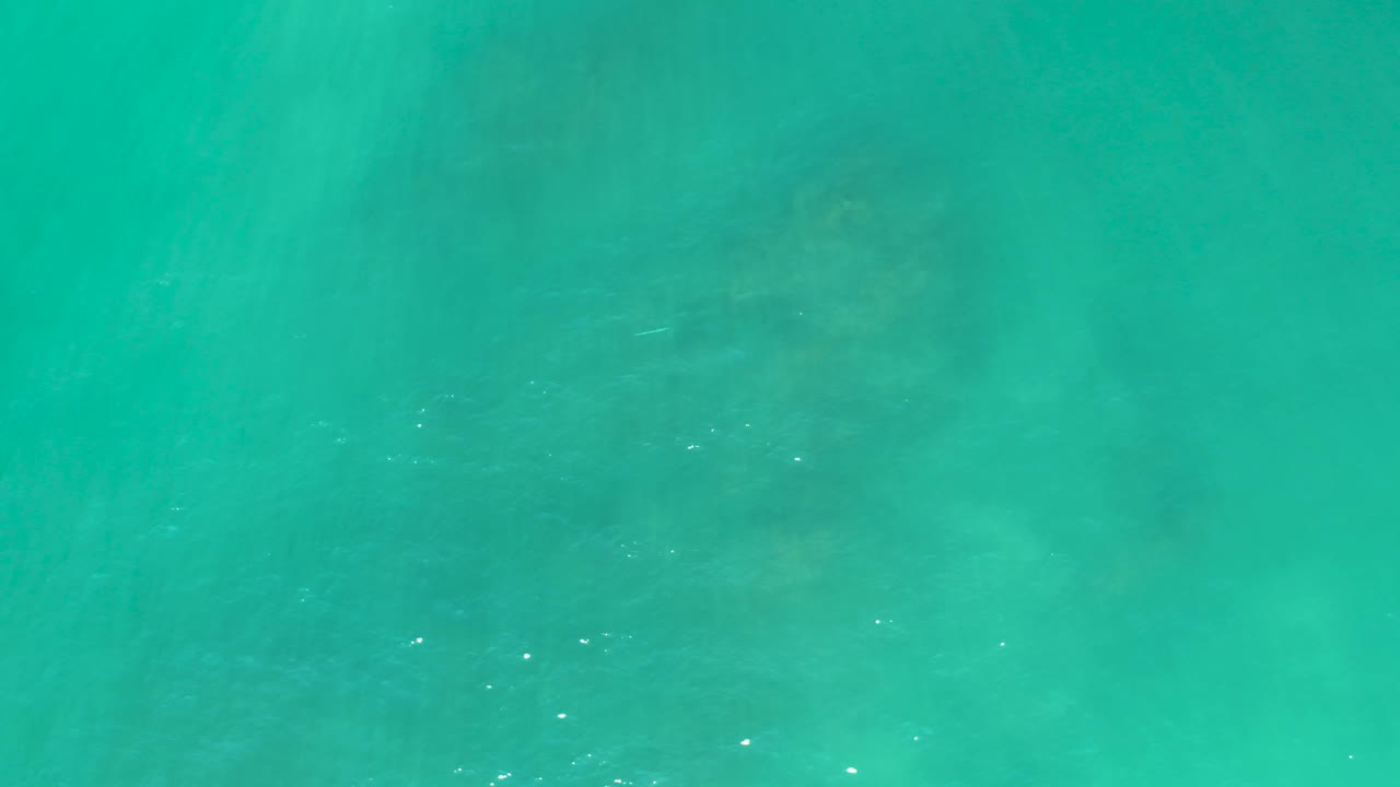 Overhead view of clear turquoise ocean water in San Luis Beach East Cape Los Cabos, with darker underwater forms more visible near the center and no visible people.