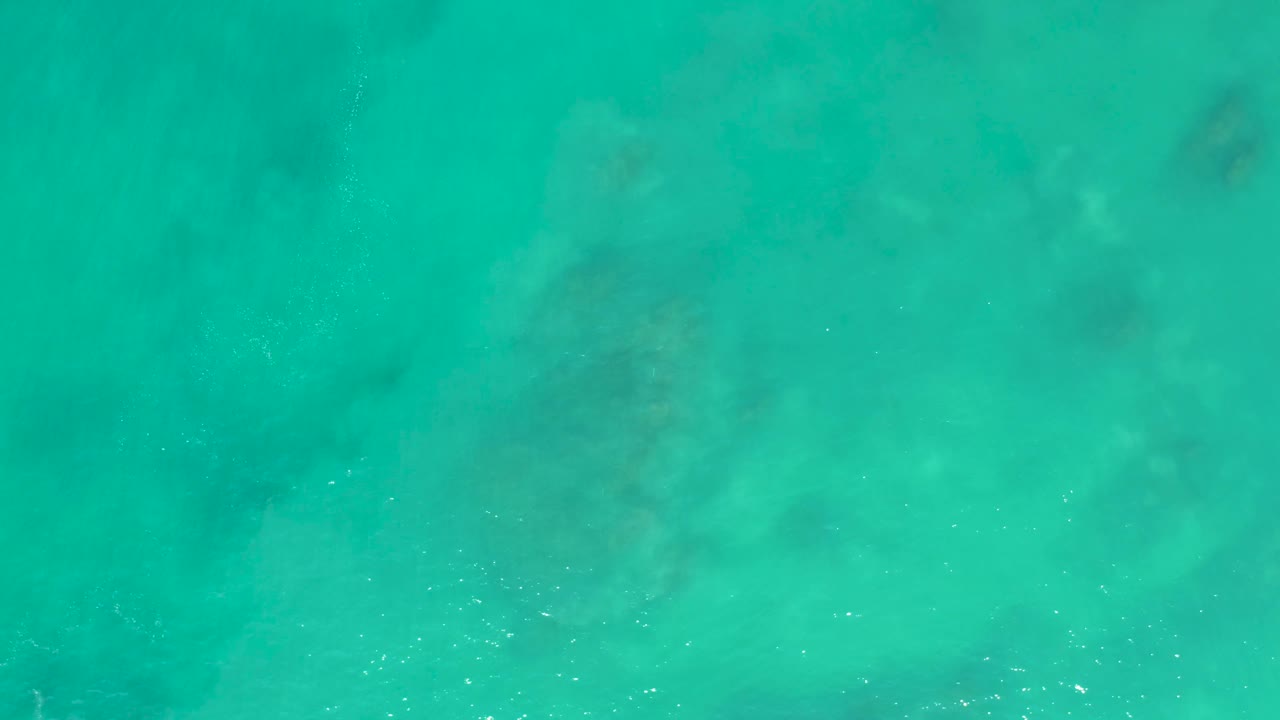 Overhead turquoise ocean view at San Luis Beach in Mexico, with darker seabed shapes under the water and light bubbling near the middle of the frame, no visible people.