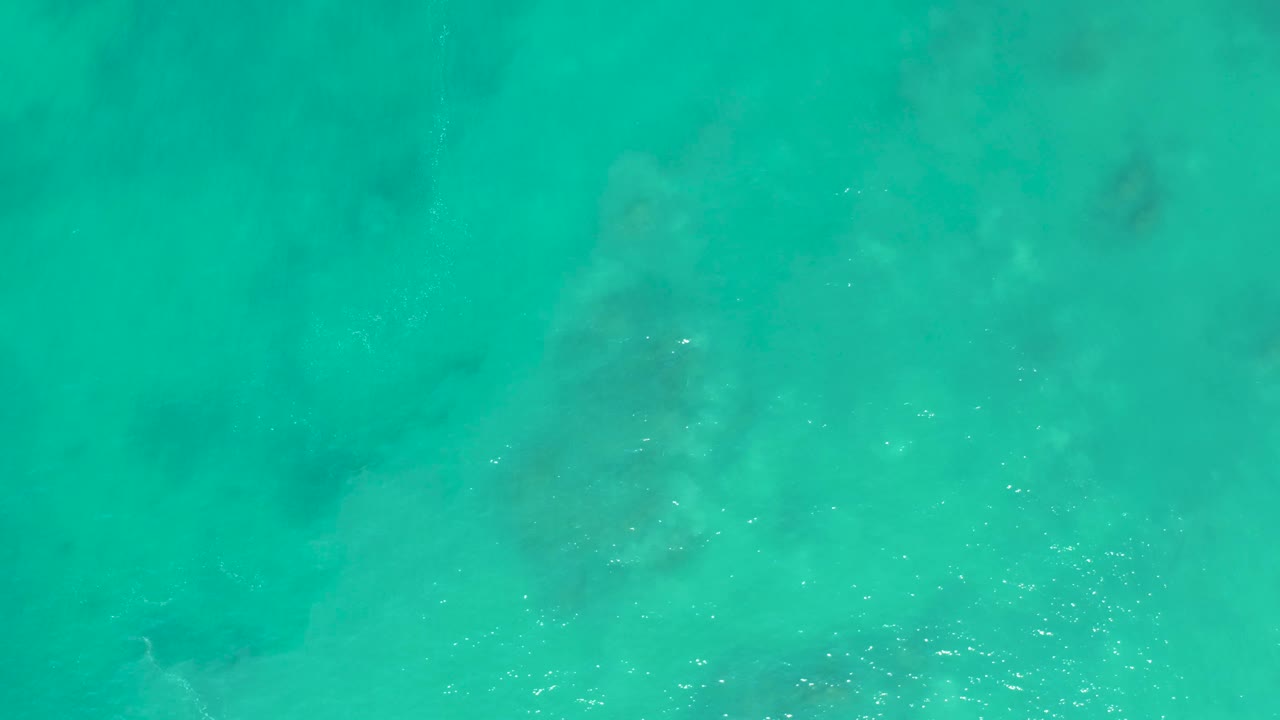 Overhead San Luis Beach water frame with clear turquoise color, a larger foam patch near the lower edge, and no visible people.