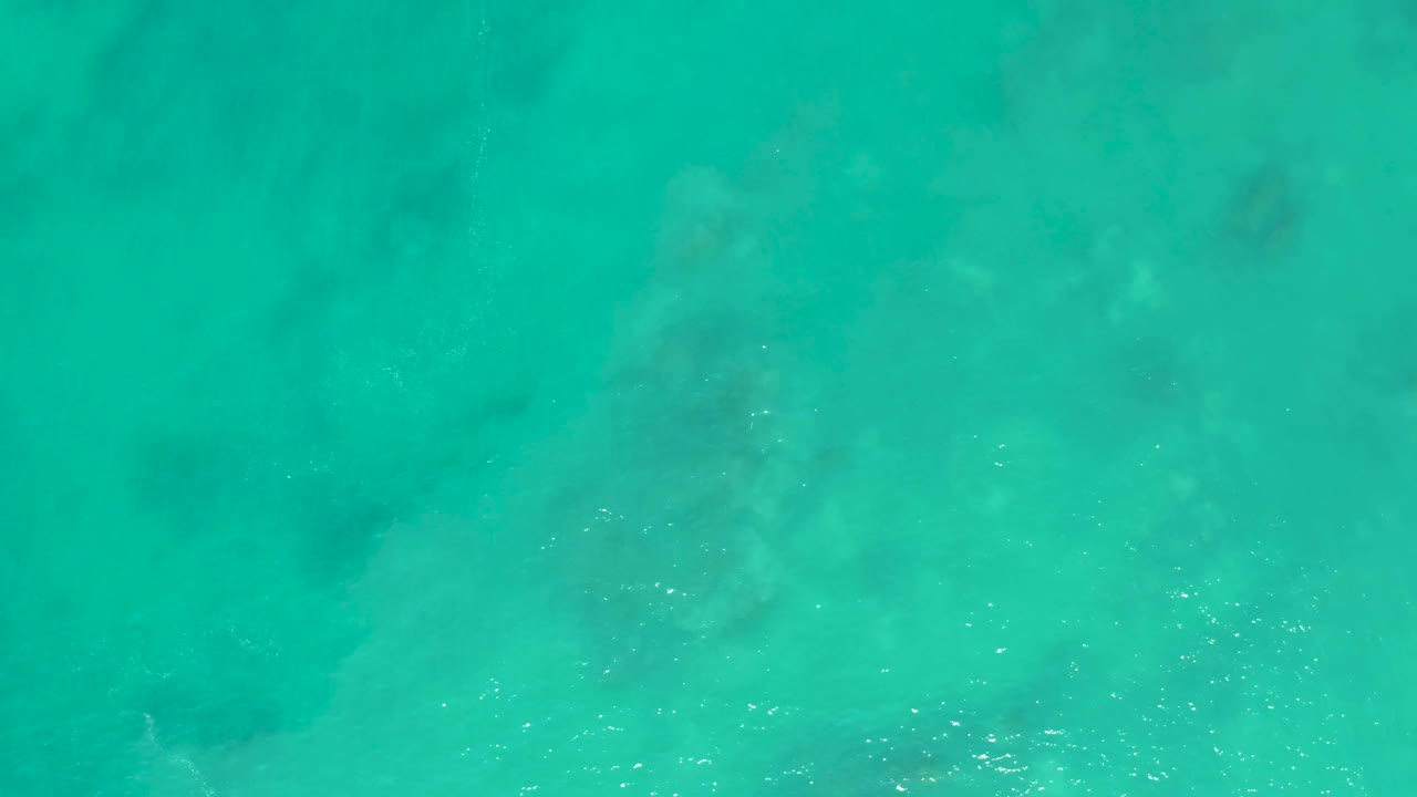 Top-down frame from San Luis Beach showing green-blue ocean water with scattered white foam and faint darker seabed areas, with no visible people.