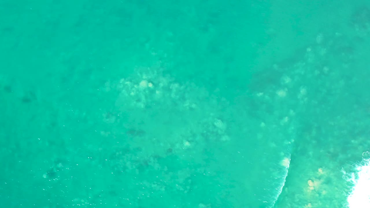 Overhead San Luis Beach water frame with green-blue shallows, soft underwater rock shapes, and a bright white patch of breaking wave foam at the right, no visible people.