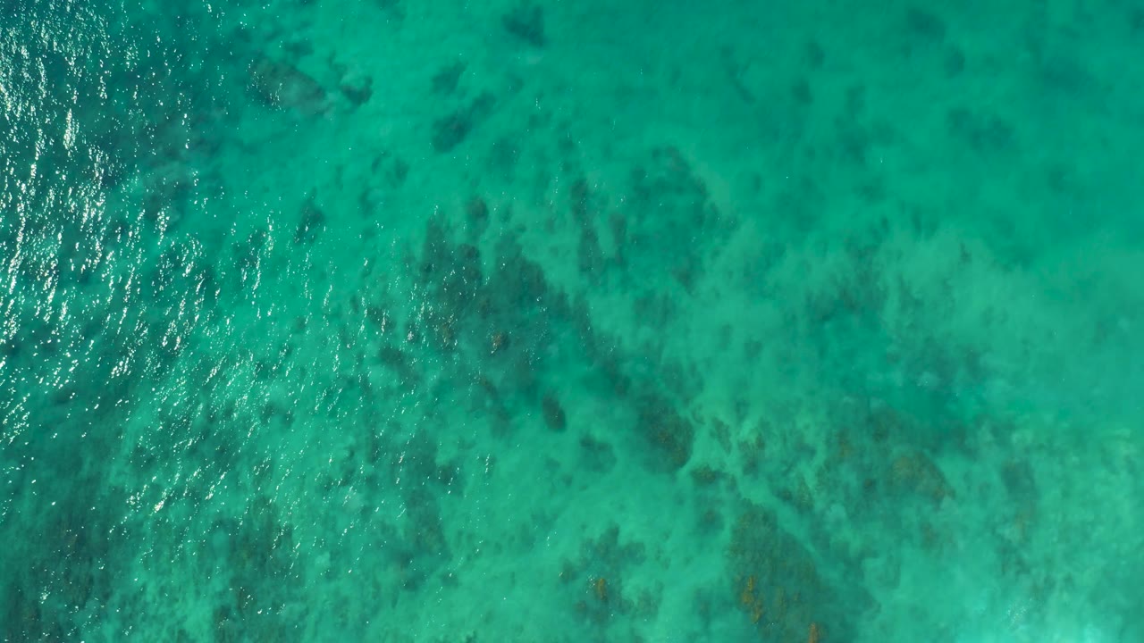 Top-down turquoise sea at San Luis Beach, East Cape, Los Cabos, Mexico, with mottled dark coral or rock patches visible beneath shifting surface reflections and no visible people.