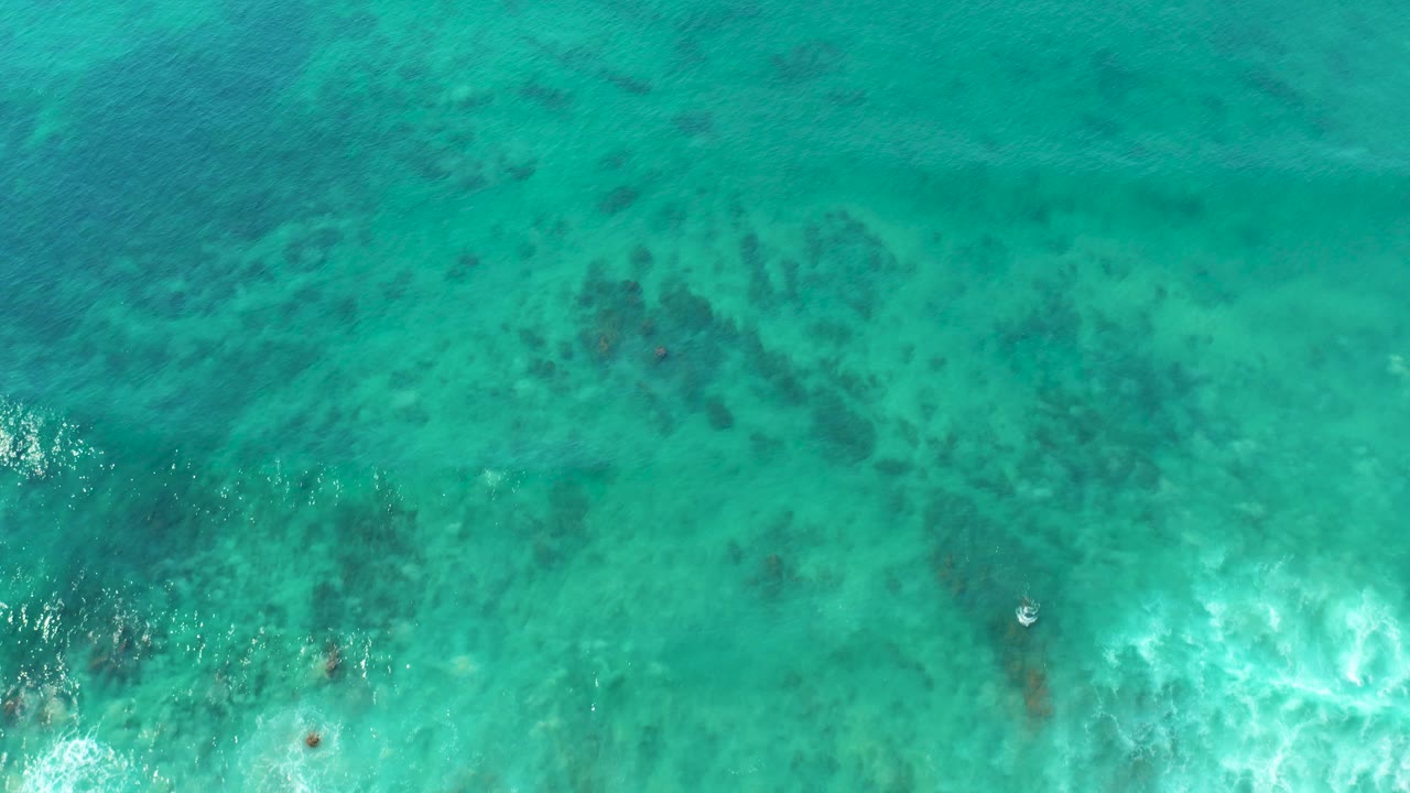 Top-down turquoise ocean water with dark reef patches and white foam at the lower edge.