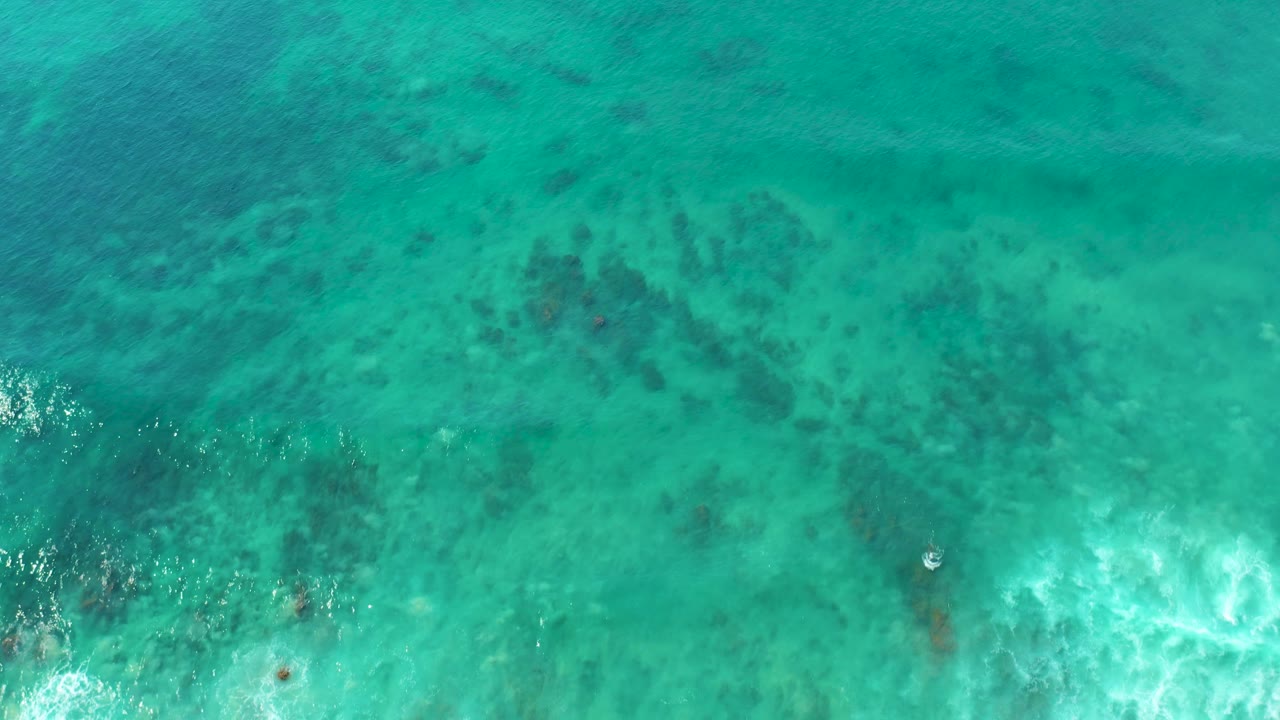 Overhead San Luis Beach water view with reef-like dark patches, a pale surface speck near the center-left, white foam lower in frame, and no visible people.