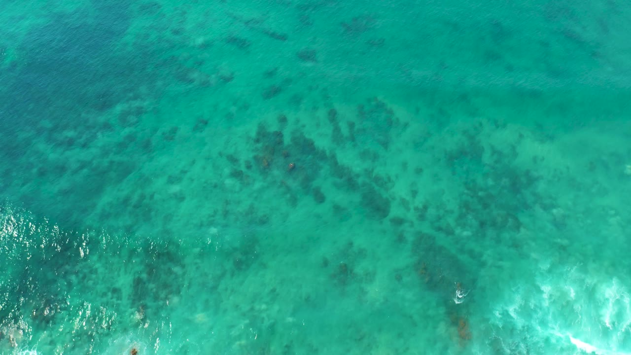 Aerial view over shallow turquoise water in the San Luis Beach clip, with dark reef texture, a band of white foam below center, sun glints, and no visible people.