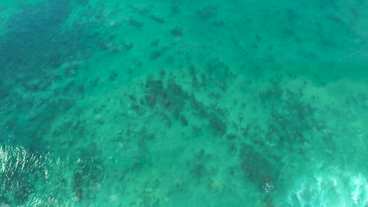 Top-down San Luis Beach water frame with turquoise surface, dark underwater coral-like patches, white foam along the lower left edge, and no visible people.