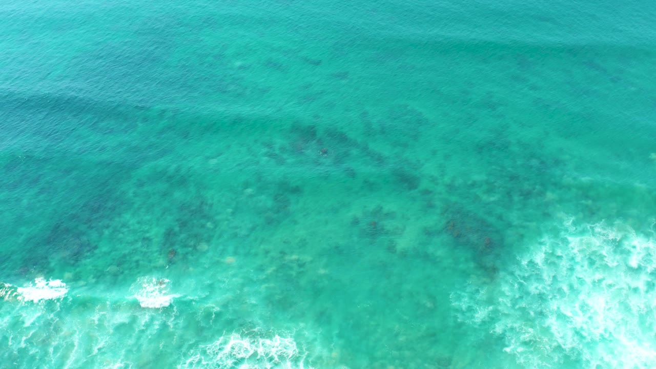 Top-down view of turquoise water at San Luis Beach East Cape Los Cabos BCS Mexico, with white foam entering from the lower edge, darker reef patches beneath the surface, and no visible people.
