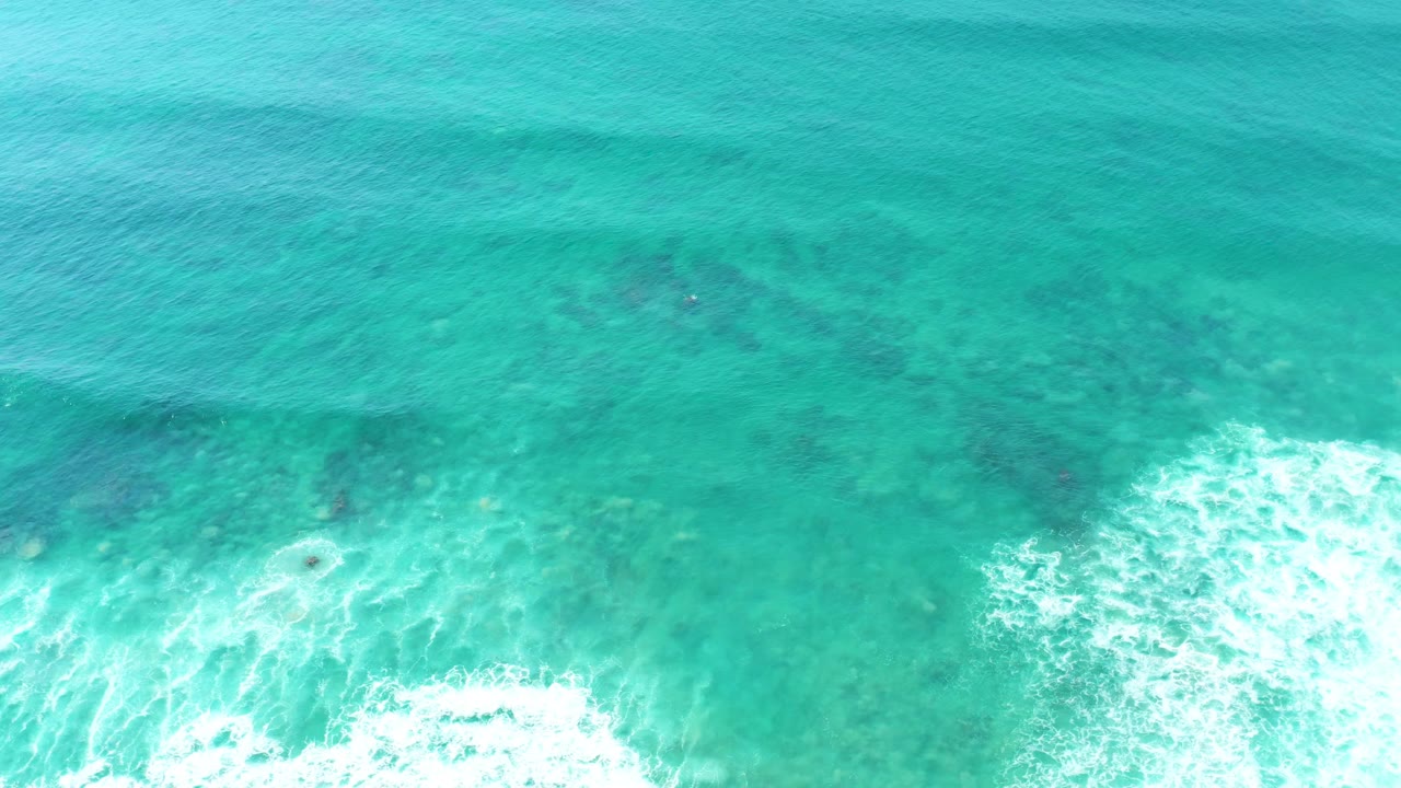 Overhead view of turquoise water at San Luis Beach East Cape Los Cabos, with dark reef patches under the surface and white foam entering along the lower edge, no visible people.
