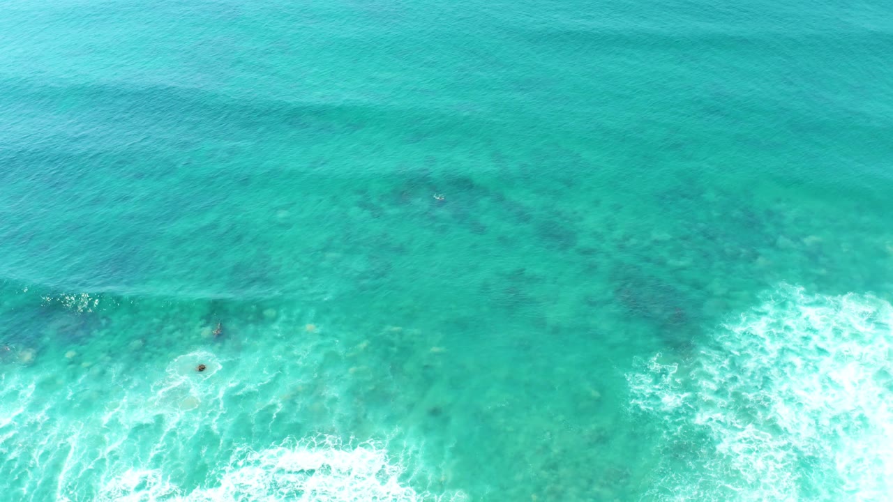 Aerial view at San Luis Beach East Cape Los Cabos with darker reef shapes beneath turquoise water and a broken white wave edge near the bottom of frame, no visible people.