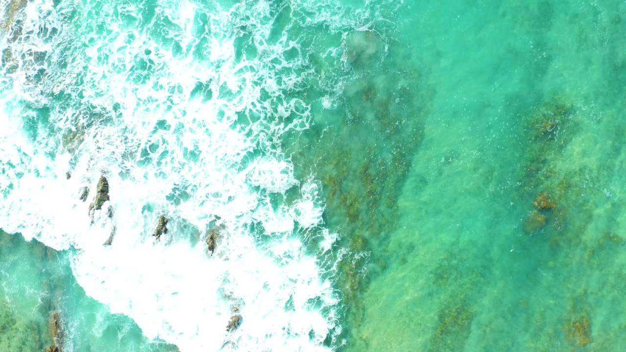 Top-down view of turquoise water at San Luis Beach, East Cape Los Cabos, with foamy surf entering from the upper left over dark rocks and no visible people.