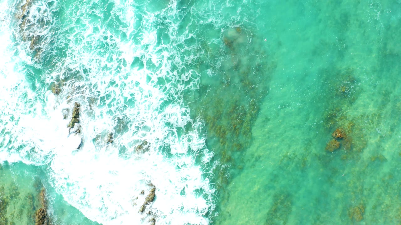 Overhead San Luis Beach water view with the main white foam mass shifting left of center and darker rock shapes visible through the turquoise water, with no visible people.