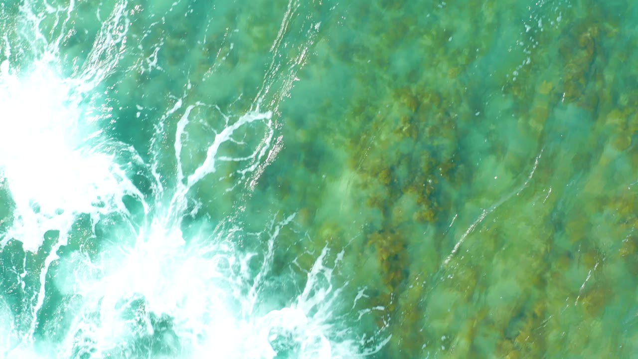 At San Luis Beach East Cape Los Cabos BCS Mexico, foamy surf remains along the left side while the rest of the overhead view shows clear turquoise water and mottled seabed, with no visible people.