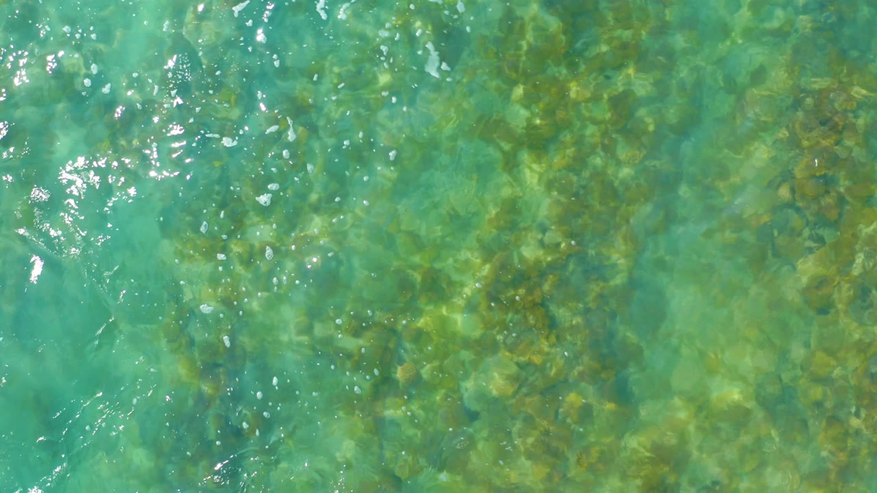Turquoise water fills the frame with submerged rocks and algae visible below brighter white ripple highlights, with no visible people.