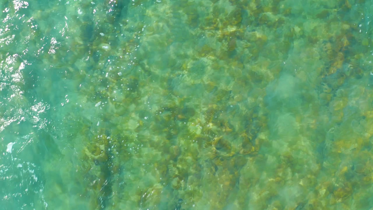 Clear green-blue ocean water fills the frame at San Luis Beach East Cape Los Cabos, with darker underwater rock patches and scattered bright surface reflections, no visible people.