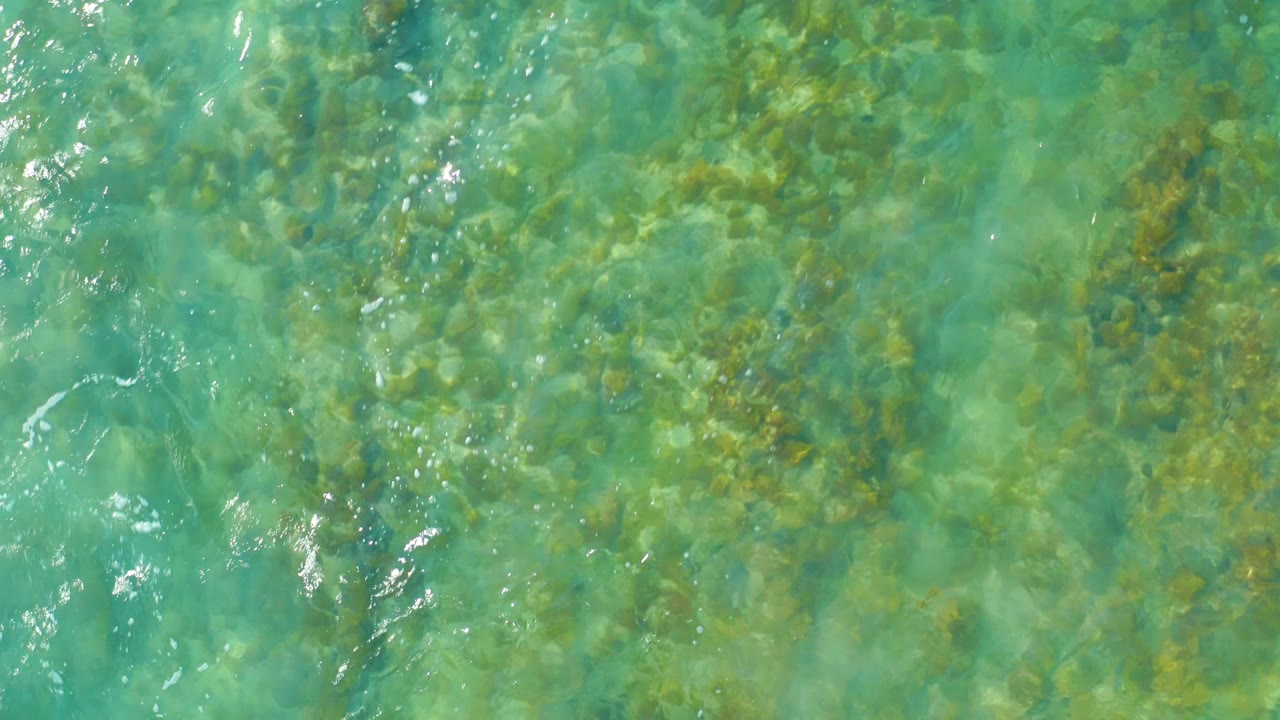 Top-down San Luis Beach East Cape Los Cabos water view with pale turquoise areas, darker submerged rocks, and small white glints on the rippled surface, no visible people.