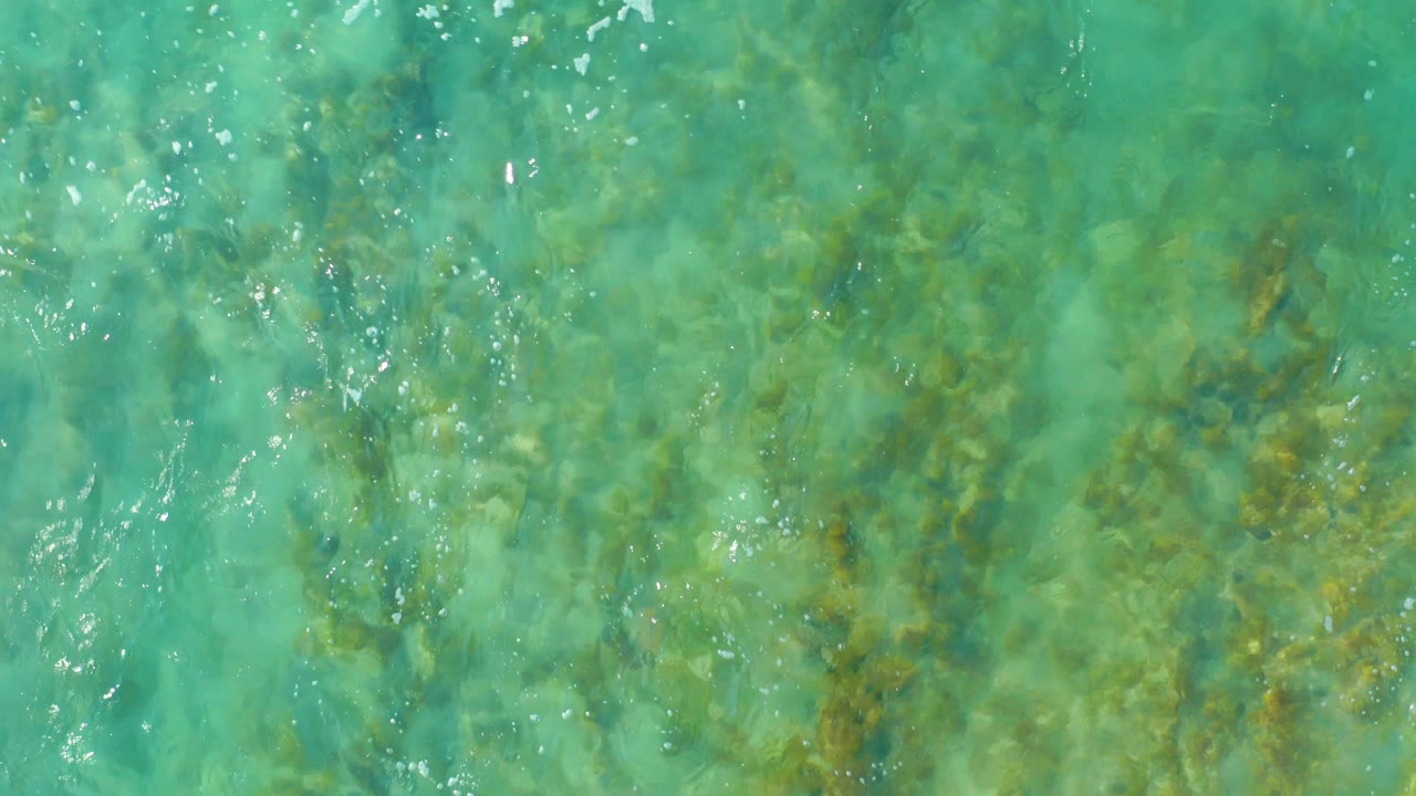 Overhead view of turquoise shallow water at San Luis Beach East Cape Los Cabos, Mexico, with submerged rocks, seaweed patches, small white reflections, and no visible people.