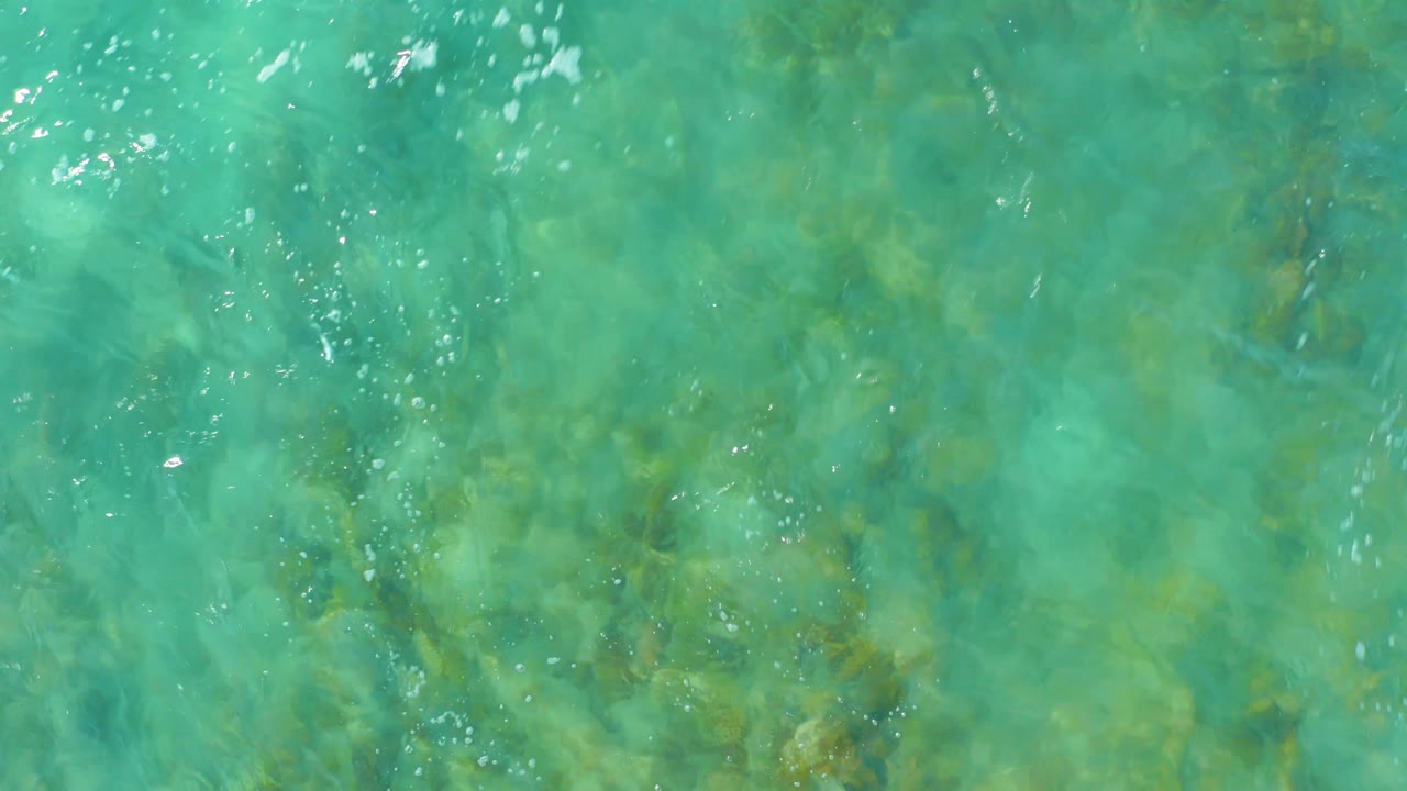 Overhead shallow ocean water at San Luis Beach East Cape Los Cabos, Mexico, shows seaweed and rock shapes beneath a turquoise surface with light ripples and no visible people.