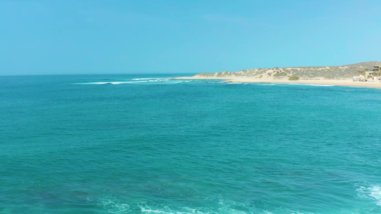 Wide turquoise ocean at San Luis Beach East Cape Los Cabos Mexico with a distant sandy shoreline and low coastal buildings, no visible people.