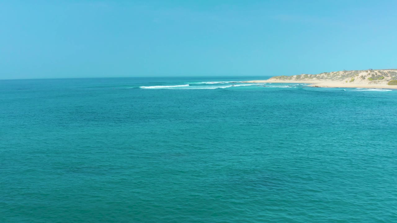 Turquoise ocean fills the foreground at San Luis Beach East Cape Los Cabos, with a thin white wave line near pale dunes and no visible people.