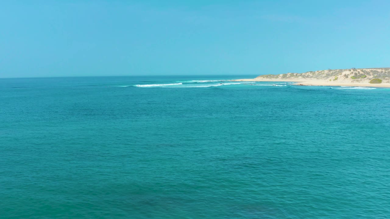 A calm turquoise ocean surface sits below a clear sky, with small breaking waves tracing the San Luis Beach shoreline and no visible people.