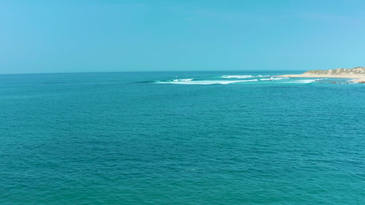 Opening frame of San Luis Beach, East Cape Los Cabos, with turquoise water in the foreground, a distant sandy shoreline, small white surf lines, blue sky, and no visible people.