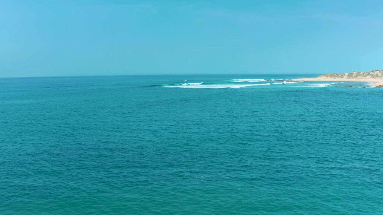 One second in, the San Luis Beach coastline remains distant while low white waves break parallel to shore across the blue-green water, with no visible people.
