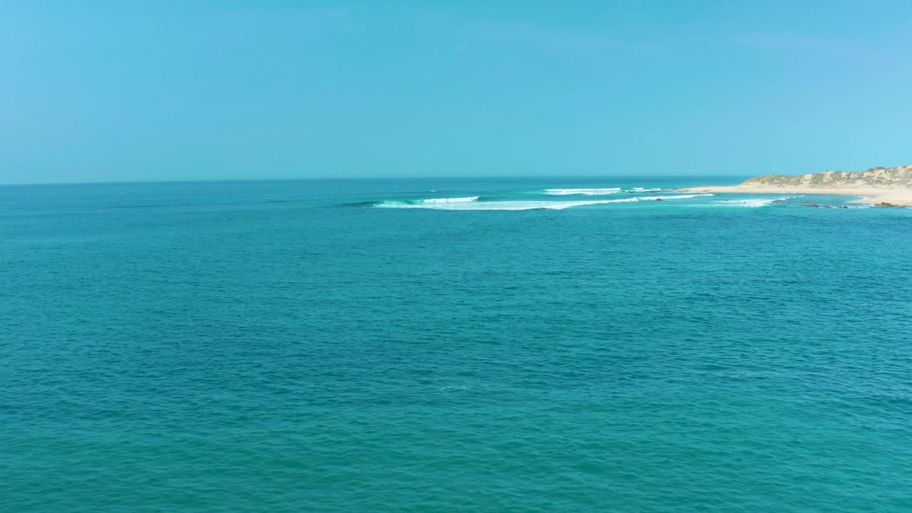 At 0:02, the frame holds a broad turquoise ocean surface, a thin horizon, and white surf breaking along the distant San Luis Beach shoreline with no visible people.