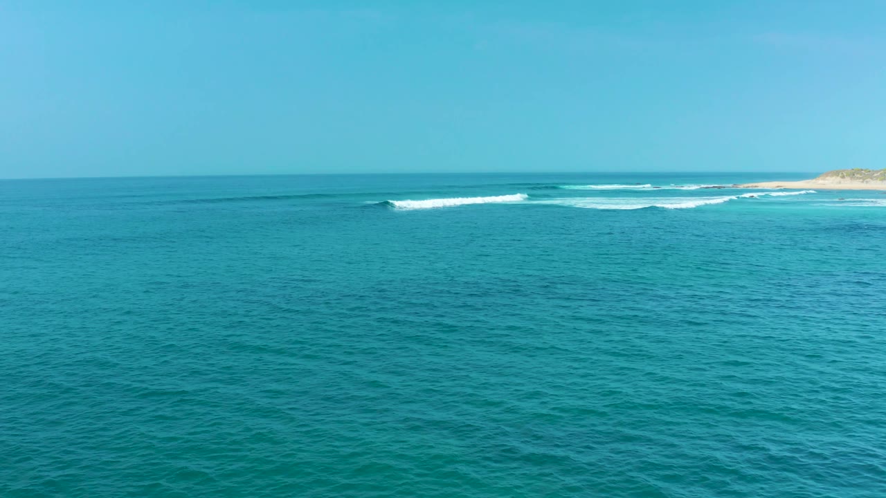 Wide coastal frame from San Luis Beach with open turquoise water in front, whitecaps breaking near the right side shoreline, clear sky, and no visible people.