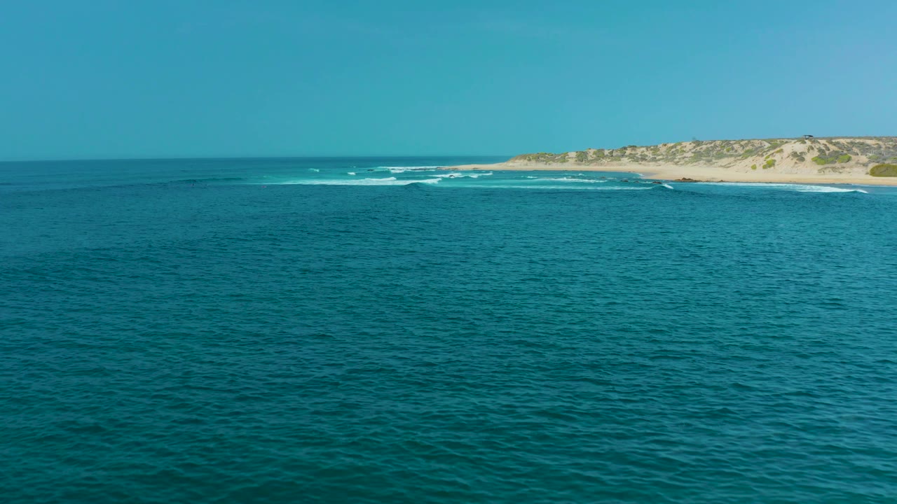 At San Luis Beach on Mexico's East Cape, blue ocean fills the foreground while white surf breaks near a sandy dune-backed shoreline, with no visible people.