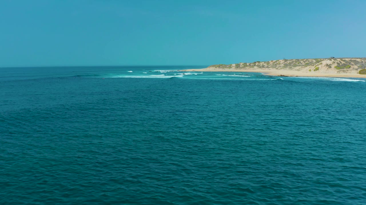 A wide view shows rippled blue water in the foreground, a band of white surf near San Luis Beach, and pale dunes under a clear sky, with no visible people.