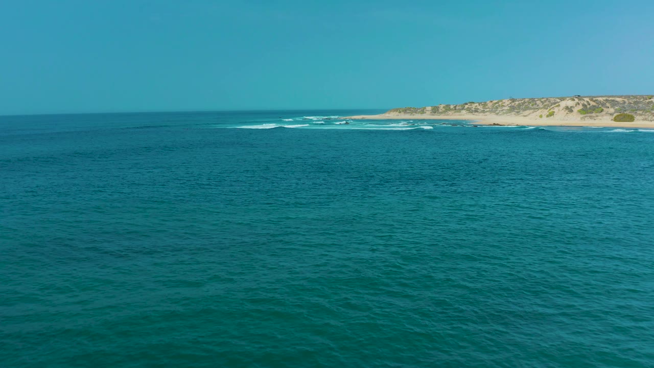 At 0:03, San Luis Beach East Cape Los Cabos appears as a distant sandy shore beyond turquoise water, with small waves breaking left of the dune line and no visible people.