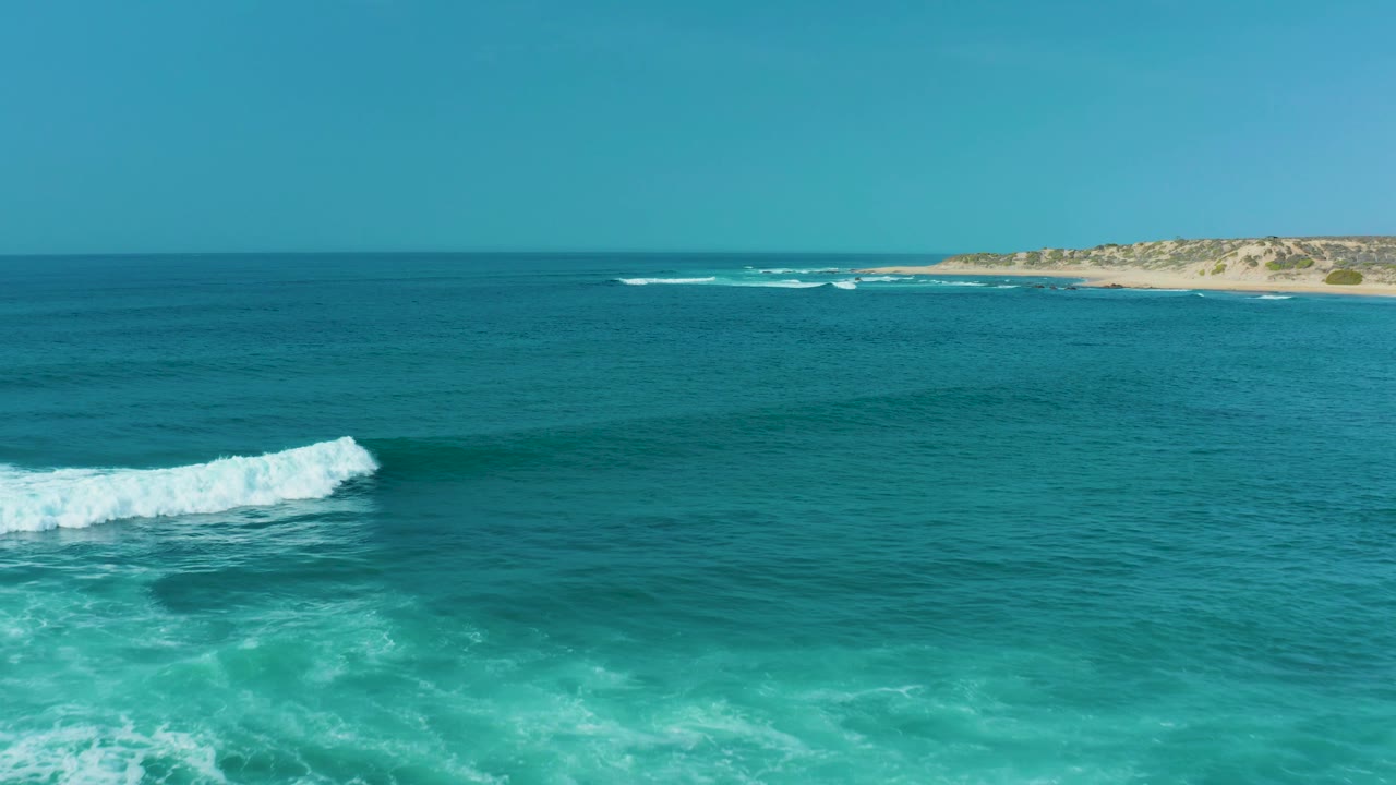 Turquoise water at San Luis Beach East Cape Los Cabos with a white wave breaking in the foreground, a sandy shoreline in the distance, clear blue sky, and no visible people.