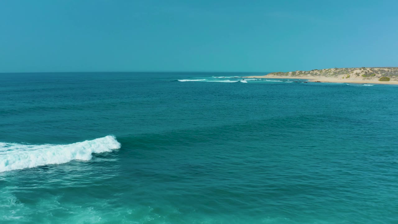 White wave crest moves across the turquoise sea at San Luis Beach East Cape Los Cabos, with open water dominating the frame, sandy shore at the top edge, and no visible people.