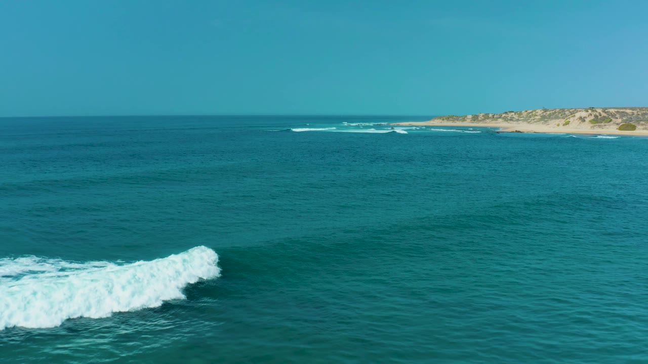 Turquoise ocean at San Luis Beach East Cape Los Cabos with a breaking wave entering from the lower left, distant sandy coastline across the background, clear sky, and no visible people.