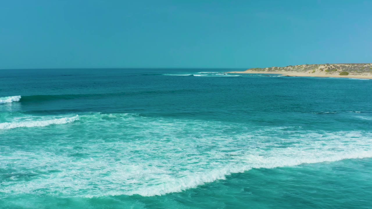 At 0:01, white surf lines spread across the lower half of the San Luis Beach frame while deep blue water and the sandy East Cape shoreline remain in the background, with no visible people.
