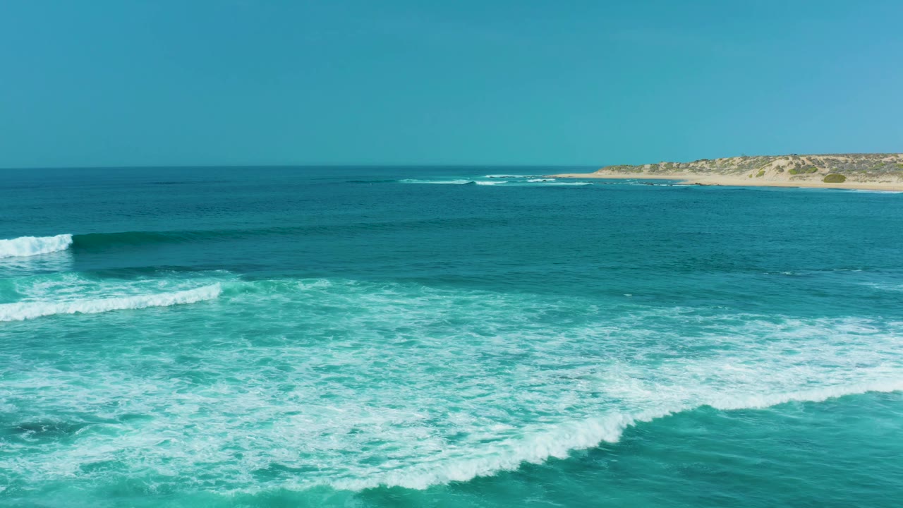 At 0:02, a brighter band of breaking surf crosses the foreground water at San Luis Beach, with a sandy point and low shoreline visible near the horizon and no visible people.