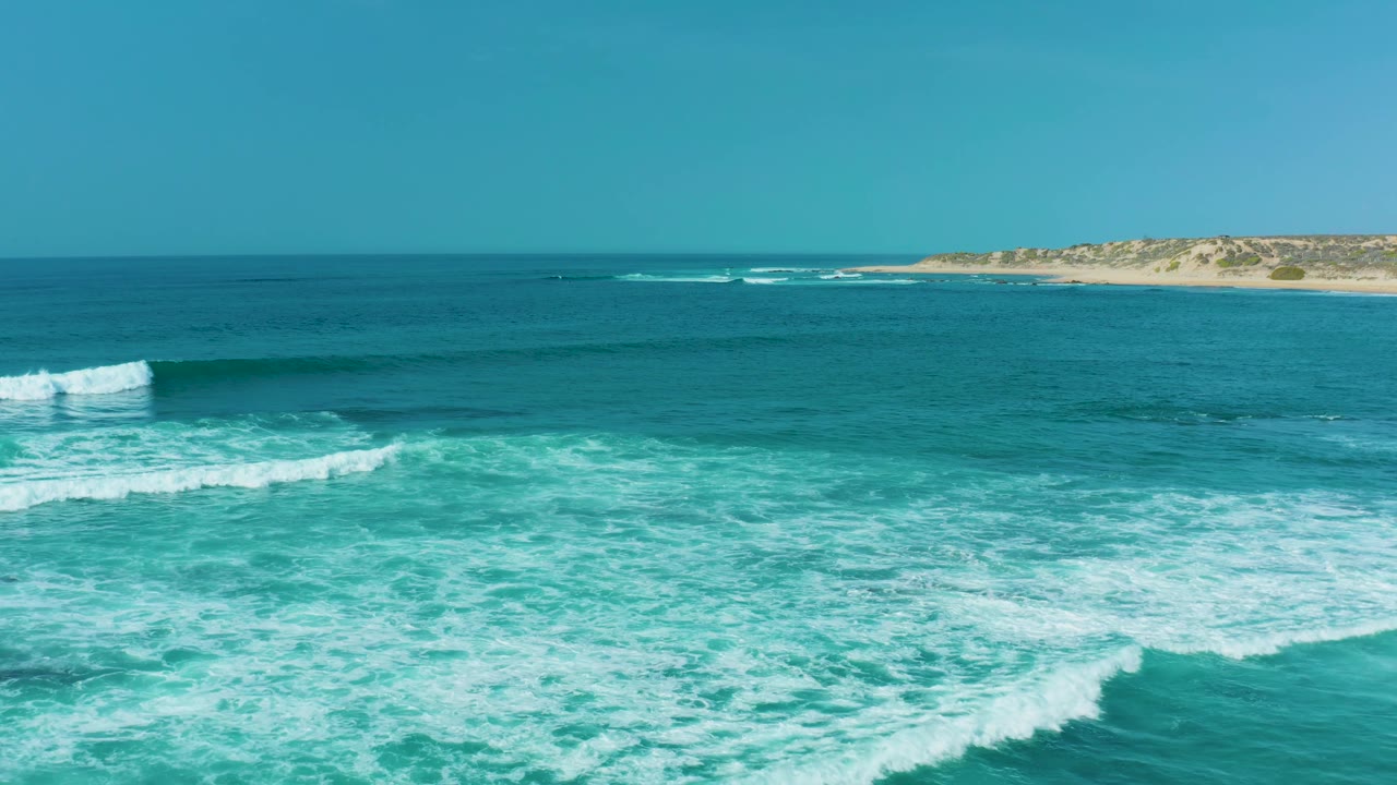 At 0:03, broad white foam sheets cover more of the foreground at San Luis Beach while the darker offshore water and distant sandy coastline stay level across the upper frame, with no visible people.