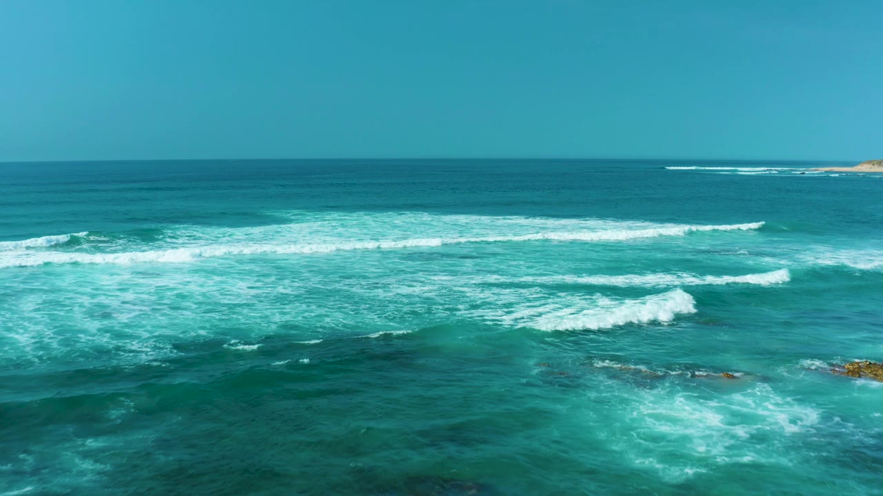 Breaking white waves spread across turquoise water at San Luis Beach, East Cape Los Cabos, with small rocks near the right side and no visible people.