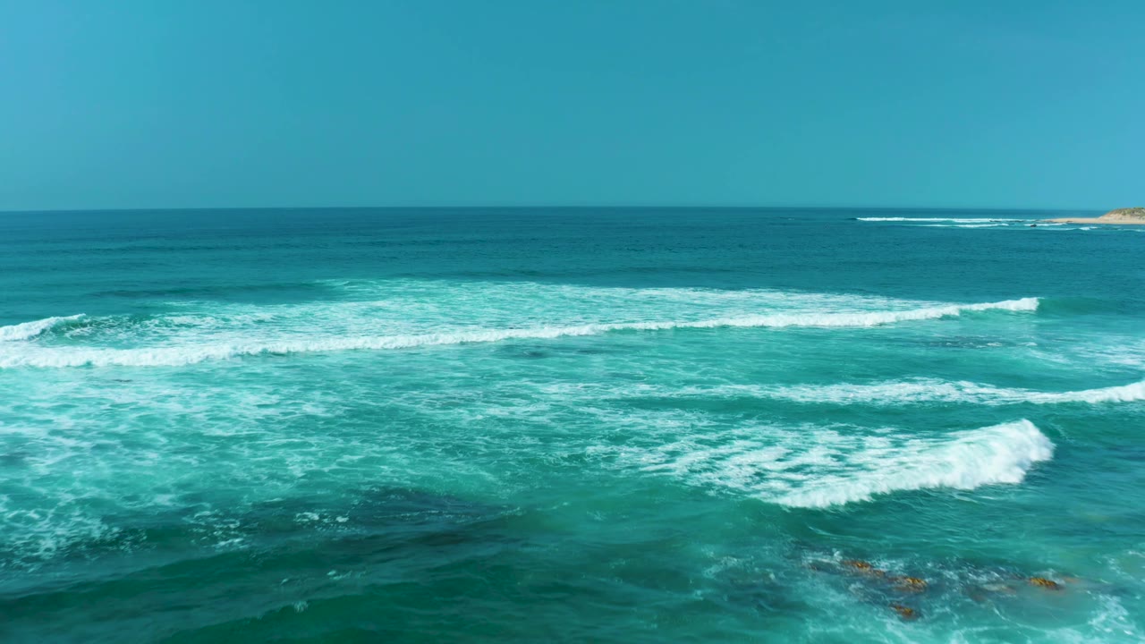 Wide view over San Luis Beach, East Cape Los Cabos water, showing white wave lines, open blue ocean, a small distant rock near the horizon, and no visible people.