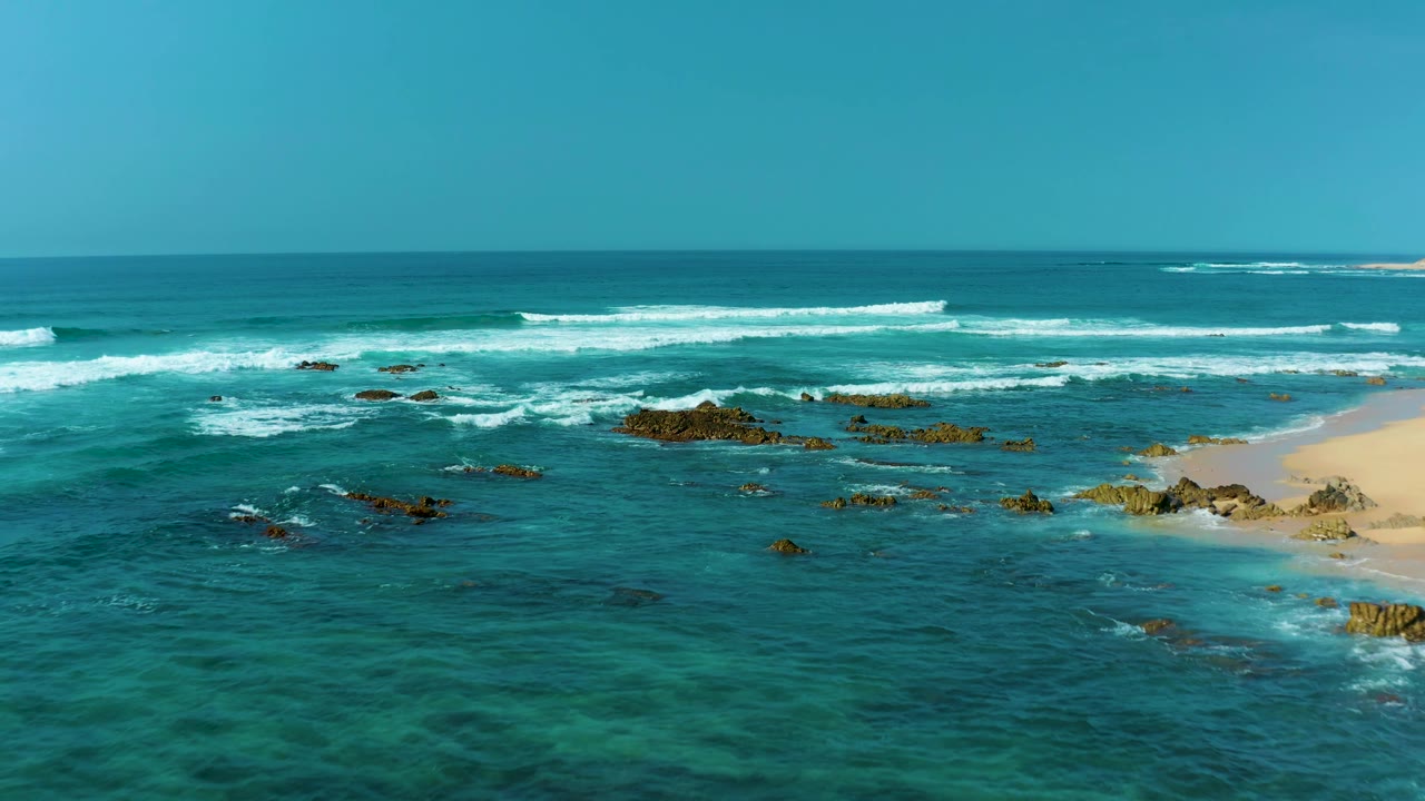 Aerial frame from San Luis Beach East Cape Los Cabos, Mexico, showing white wave bands crossing shallow turquoise water toward exposed rock clusters, with no visible people.