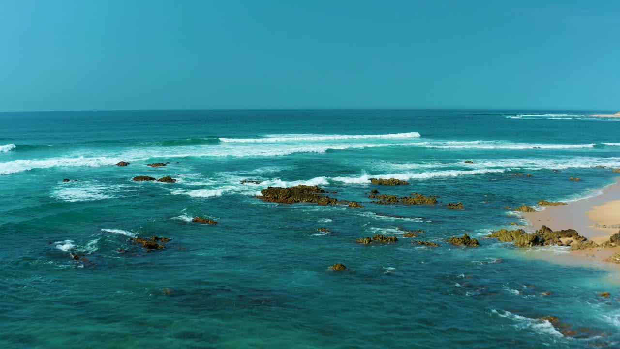 San Luis Beach East Cape Los Cabos, Mexico, appears from above with foam wrapping around dark rock outcrops, teal water across the foreground, sand at the right edge, and no visible people.